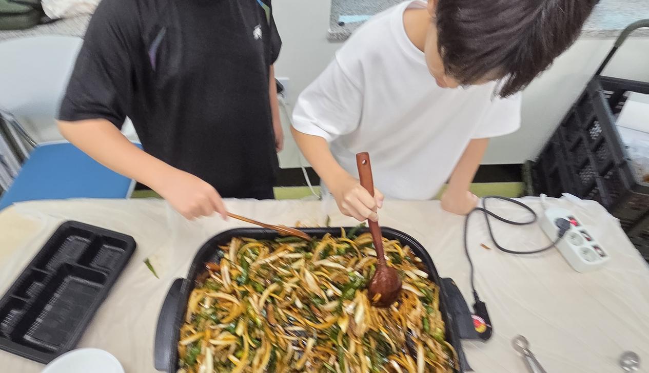 나무 젓가락과 나무 숟가락을 든 두 아이가 고추잡채를 볶으며 즐거워하는 모습